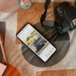 Điện thoại Android hiển thị ứng dụng Photoshop trên giường cạnh máy ảnh và sổ tay