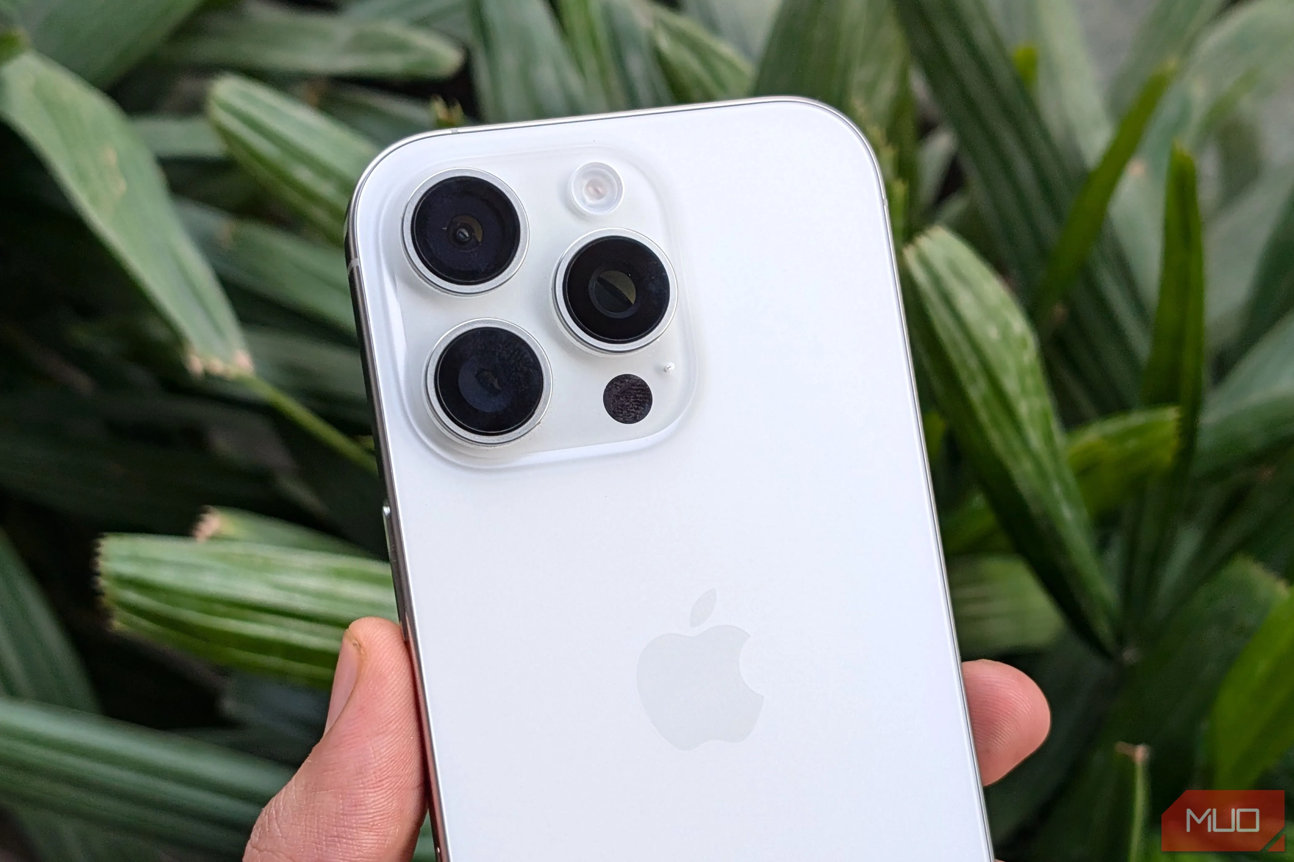 iPhone 16 Pro màu trắng được cầm trên tay, phía sau là cây xanh, thể hiện thiết kế cao cấp và camera nổi bật của điện thoại.