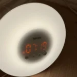 Một chiếc đồng hồ báo thức bình minh với ánh sáng dịu nhẹ đặt trên sàn, minh họa thiết bị hỗ trợ giấc ngủ tự nhiên.