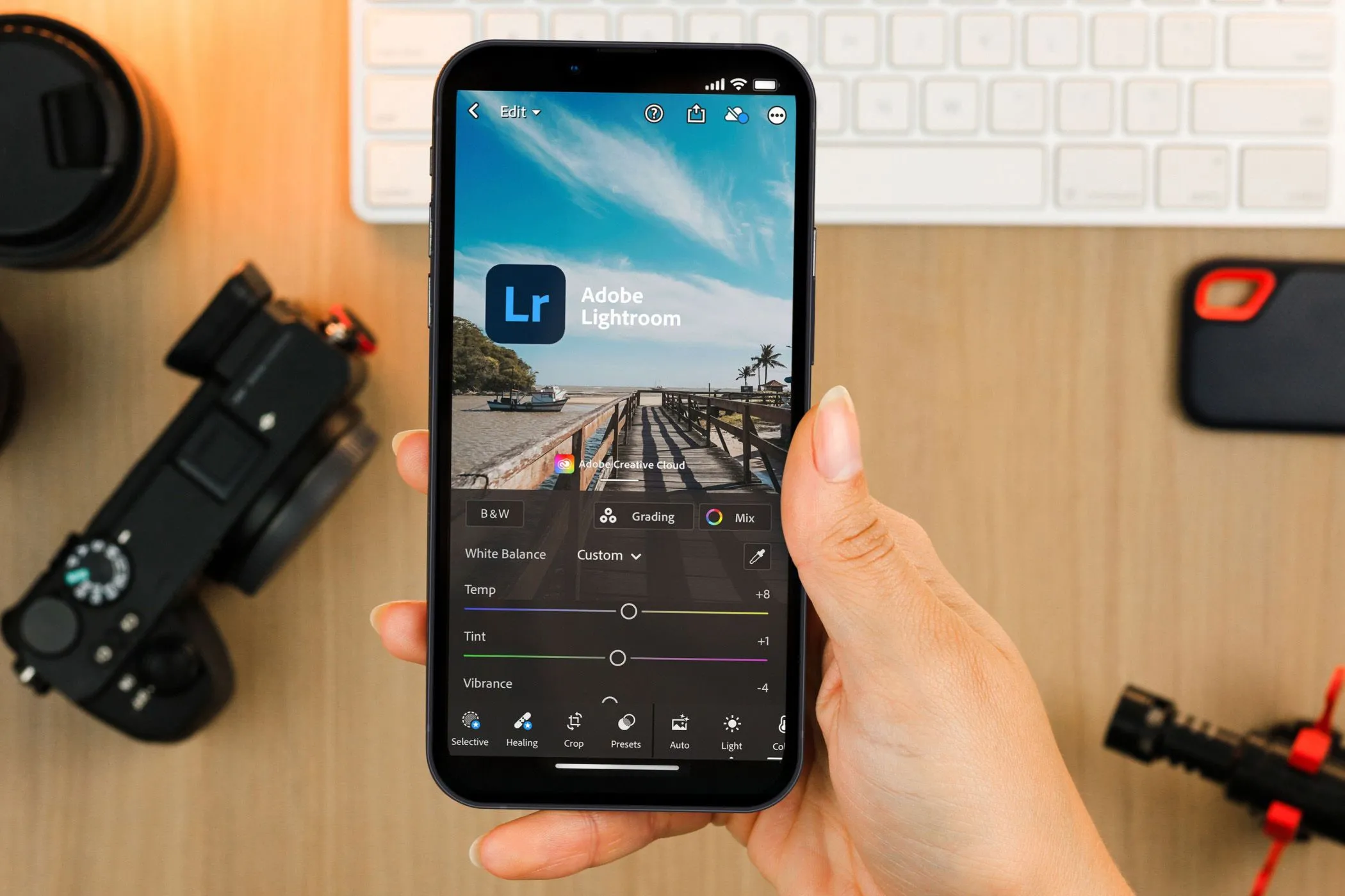 Người dùng đang cầm smartphone hiển thị giao diện ứng dụng Lightroom Mobile, có thiết bị máy ảnh phía sau